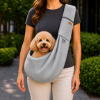 Gray crossbody pet sling carrier worn outdoors with a small curly coated dog inside.